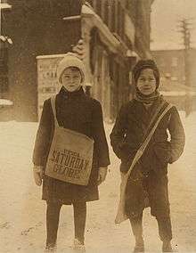 Two young newsboys smiling and standing in the snow. One boy is holding a bag.