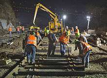 A crew of men in orange vests with reflector stripes and neon green hard hats over their clothing laying new railroad ties. Behind them is a large yellow piece of heavy equipment. It is night, and the scene is illuminated by large lights.