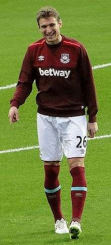 A smiling man with light coloured hair and wearing a claret football training jersey, white shorts, claret socks and white football boots walks across a football pitch face-on to the camera.