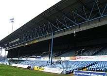 An imposing colour shot of a traditional British-style grandstand, mostly painted blue and filled with blue seats. A sign around its centre, hanging from the ceiling, reads "Cardiff City F.C.".