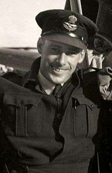 Half-length informal portrait of grinning man in dark-coloured battle jacket, wearing peaked cap