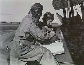 Two men in flying suits, one in the cockpit of a biplane and the other standing beside him