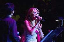 Woman singing on stage in pink lighting, with man next to her