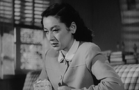 Noriko, wearing the same dress as in the Noh sequence, is shown seated on a sofa from a slightly lower angle; a window and some books are dimly visible in the background.