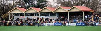 A grassed sporting oval with an old stand with a yellow and red roof, filled with spectators