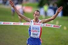 A young man representing France lifts his arms as he breaks the tape at the finishing line of a race.