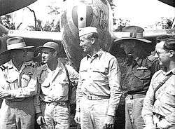 Five men in light-coloured uniforms standing in front of a military aircraft