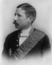 Photo of mustachioed man, in white tie and with a sash.