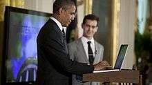 Barack Obama in a blue suit typing on computer at a podium while a white man in a light grey suit looks on