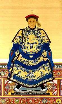 Full-face painted portrait of a severe-looking sitting man wearing a black-and-red round cap adorned with a peacock feather and dressed in dark blue robes decorated with four-clawed golden dragons.