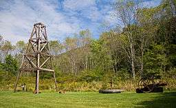A wooden derrick at left on grass, with trees and a mountain ridge in the background