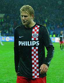 A photograph of a blonde footballer wearing a black shirt with a stripe of white and red checkers on the left side of his chest and red shorts. The man is seen walking football field with his view downwards.