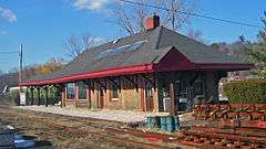 A brick building seen from its right front. It has a peaked black roof with red trim and a broad overhang. In front of it are rusted railroad tracks. Two old green passenger cars are behind it to the left.