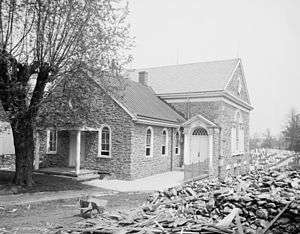 A three-quarters view of a brick church in a black-and-white photo. A barren tree is to the left and a pile of rubble lies to the right.