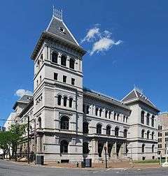 An ornate grey stone building with many arches along its front and two peaked roofs seen from across an intersection.