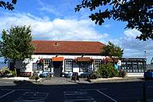 A colour picture of a pub. It has an orange pantile roof and the walls are white. There are flowers and a tree in front of it. A conservatory is on the right of the pub and the sea is visible behind it.