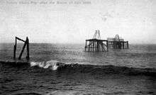 A black-and-white image of the pier after being destroyed by a storm. The deck and the chains are missing, and the nearest platform has been removed apart from a few broken piles.