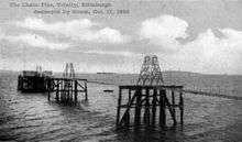 An old black-and-white photograph of a suspension pier. On the furthest platform of the pier are two buildings. There are two steamships in the background. In the foreground a rectangular object floats in the sea.