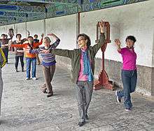 Six women, some elderly, standing in a column (except for one on the right) next to a white wall on a stone floor. All are clad in street clothes, with their left arm curled over their heads and their right arm extended.