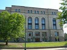 A beige five-story municipal building with large, arched windows. A tree is in front of the building.