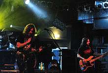 Two men with long hair are on a stage. One plays a shiny guitar and the other plays a bass guitar with a wood finish