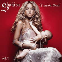 An image of a sitting woman with long blonde hair. She is wearing a sleeveless white dress that extends past her legs. She is holding an unclothed infant in her arms, who is reaching at the necklace she is wearing. The terms "Shakira" and "Fijación Oral" are respectively positioned in the top left and right corners of the image, while the term "Vol. 1" is placed in the bottom left corner.