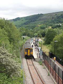 Aerial view of four carriage train, on single curved track, at station. Passengers wait to board. Lush wooded mountains make up the background on all sides. To the right, foreground, a walkway leads down to the platform.