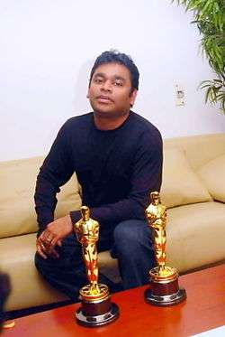 A color photograph of a man sitting on a sofa, with two golden statuettes on the table in front of him.