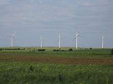  An area of flat green fields with five starkly white wind turbines standing out from the background of a blue sky.