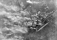 A British bomber aircraft flies over a heavily bombed agricultural landscape with many craters and plumes of smoke visible immediately below