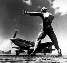 Black and white photograph of a man wearing military uniform facing a World War II-era single-engined fighter while gesturing to his left