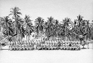 Large crowd of military men seated in front of three single-engined aircraft with palm trees in the background