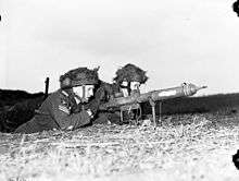 Two men in military uniform lie on a grassy bank, handling a tube shaped weapon