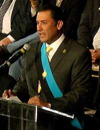 A black suited man talking to an audience in front of a microphone