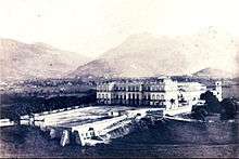 Photograph of a three-storied, neoclassical palace facade, fronted by a large open space supported by massive retaining walls and with high mountains rising in the background