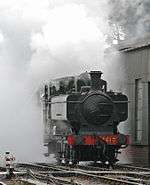 A green pannier tank locomotive appears from a cloud of its own steam and smoke.