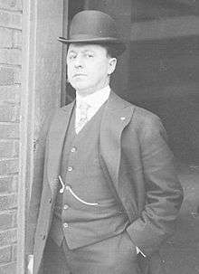 Medium balck and white shot of white man standing in a doorway wearing a dark suit and derby hat