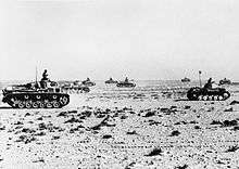 Several tanks advance to the right across the brush-covered desert.