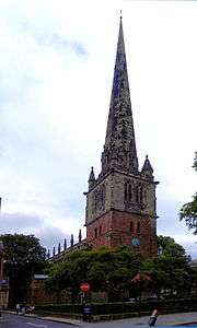 The tower of a church with a tall spire. The lower part of the tower is in red sandstone, the upper part and the spire are in a grey stone