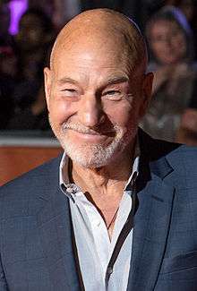 A bald caucasian man at a film premiere with a closely cropped grey beard.