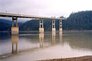 A beige bridge on supports spanning a brown colored river and evergreen hills in the background.