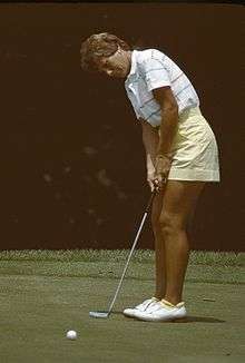 A blonde-haired woman in a mostly white shirt and yellow shorts is hitting a putt with a putter with white golfing shoes on