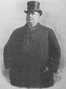 A large man stands wearing a full length coat and top hat.