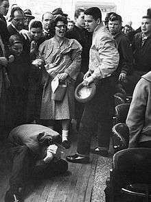 A man is curled up on the floor protecting his head with his hands while his attacker hovers over him and a crowd of bystanders watch.