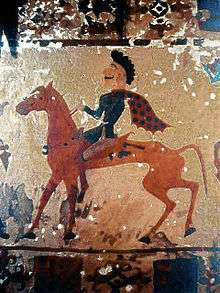 This picture is of a Pazyryk man riding a red horse shown in profile view. The man has black hair and is wearing a flowing red and blue polka dotted cape.