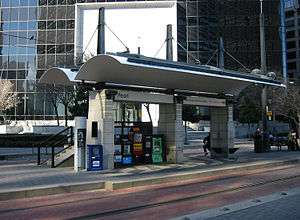Island platform station and canopy in the median of the street with only a few passengers present.