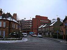 A tall red brick building towers over streets of two-storey houses. The roofs of the houses and the surrounding pavements are covered in snow