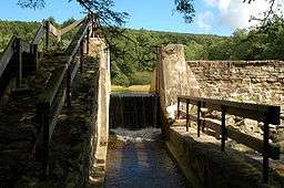 Water pours over a narrow spillway with a sunlit stone dam structure on either side