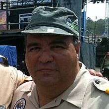 A Hispanic male wearing a beige colored button-up shirt and green cap posing for a picture outdoors.