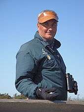 A man wearing binoculars, a brown leather visor, blue jacket, and black gloves stands facing right but with his smiling face towards the camera. He is holding a clicker counter in one hand.